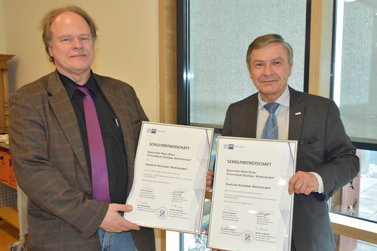 Direktor Werner Epp (links), und Werner Ehrmanntraut, Personalleiter beim BRK Kreisverband Ostallgäu halten die eben unterzeichneten Urkunden der Partnerschaft zwischen Schule und Wohlfahrtsunternehmen in Händen.